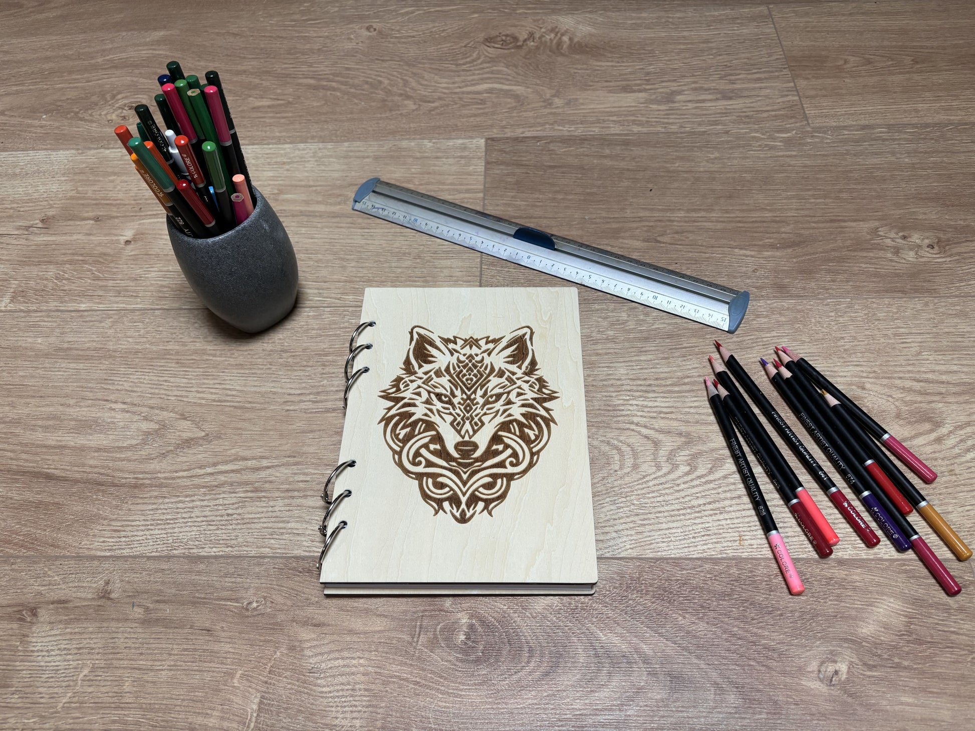 Cahier A5 avec couverture en bois (tilleul) et tenu par six anneaux en métal. Motif tête de loup. Le cahier est entouré de crayon, d'un pot à crayon et d'une règle en métal.